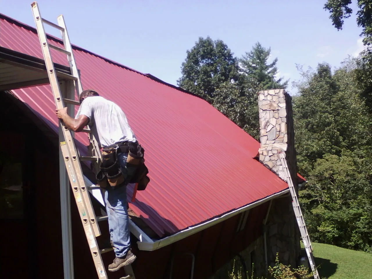 Roofer on a ladder beside a red metal roof and stone chimney — Soffit & Fascia Repair by Roofing Brothers in Carroll